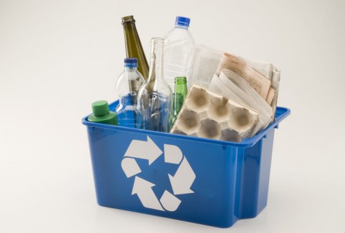 A blue plastic recycling bin with a white recycling symbol on its side, filled with various waste items including clear glass bottles, a brown glass bottle, a plastic water bottle with a blue cap, a carton of egg shells, and newspapers. The bottles are positioned upright, with some of the newspapers and paper packaging leaning against the bottles inside the bin. The background is plain white, emphasizing the contents of the bin and its clean, smooth finish, typical of professional rubbish removal services in Hounslow. The collection of recyclable materials suggests an organized approach to waste management often provided by companies like Office Clearance Hounslow, operating within the Hounslow postcode area, for efficient rubbish disposal and recycling solutions.