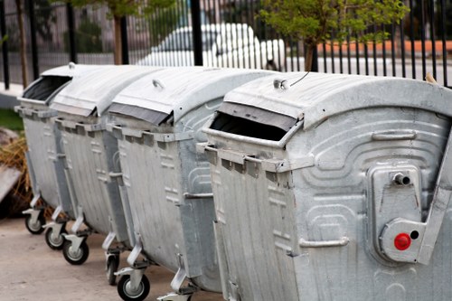 A row of four large, industrial-grade roll-front metal rubbish bins with hinged lids, situated on a paved outdoor area. The bins are made of galvanized steel, displaying a silver-grey finish with visible ridges and seams. Each bin is mounted on four small wheels for mobility, with the front wheels showing some wear. The lids are slightly raised, revealing dark interior openings for waste insertion. The background features a black metal fence with vertical bars, behind which are parked white vehicles and some trees, indicating a suburban or urban environment. The scene is lit with natural daylight, suggesting a typical daytime setting. This arrangement of bins is indicative of a commercial or residential waste collection setup, aligning with rubbish removal services such as those offered by Office Clearance Hounslow, especially in the Hounslow area of west London.