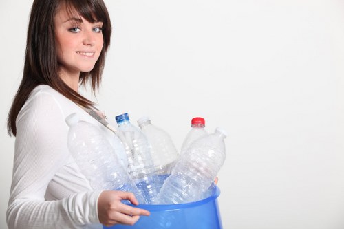 A young woman with shoulder-length brown hair and a light complexion is standing against a plain white background, holding a blue plastic bin filled with several clear plastic bottles. The bottles vary in size and shape, with some having red or grey caps, and are partially filled with liquid, indicating they are used or recycled. She is wearing a white long-sleeved top and has a neutral expression, looking slightly to her left. The scene suggests a focus on waste collection or recycling activities, relevant to rubbish removal services in Hounslow. The setting appears to be indoors with professional lighting, emphasizing the clean and organized appearance of the waste management process. This image visually supports topics related to recycling, rubbish disposal, and environmentally responsible waste management in the local area of Hounslow or nearby postcodes, highlighting efforts to promote sustainability and proper waste handling.