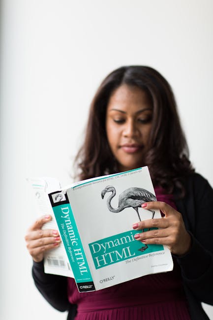 A woman with medium to dark skin tone and long, dark hair is standing indoors against a plain white wall, holding an open book titled 'Dynamic HTML' with both hands. She appears to be reading or exami