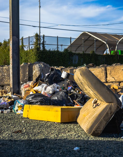 A collection of discarded black rubber tires lying on a grassy area next to a line of leafless and leafy young trees. The tires vary in size, with some stacked and others scattered individually on the