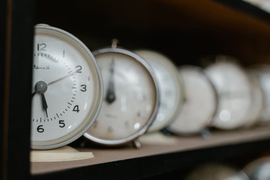 A collection of various analog and digital alarm clocks stacked together on a metal shelf or storage area. The clocks are made from materials such as plastic, metal, and glass, with textures ranging f