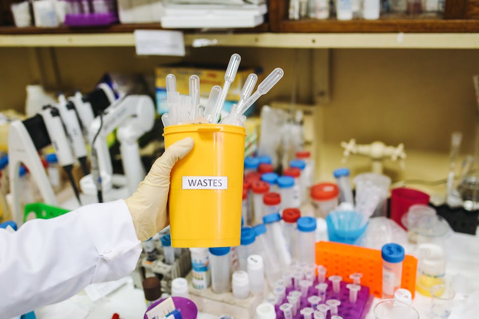 A person's hand, clad in a grey sleeve, is seen inserting a used lithium battery, with a yellow and blue label marked 'VARTA' and 'D , into a green container designed for battery recycling. The contai