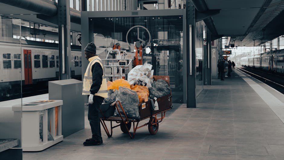 A middle-aged man with dark hair, wearing a black t-shirt with white and grey text and dark trousers, is seen disposing of rubbish into a stainless steel, cylindrical public waste bin located on a pav